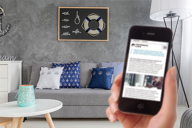 A right hand holding a phone with Greenberg LASIK website on the screen and a living room backdrop with couch and pillows. The phone is out of focus while the backdrop is in focus.