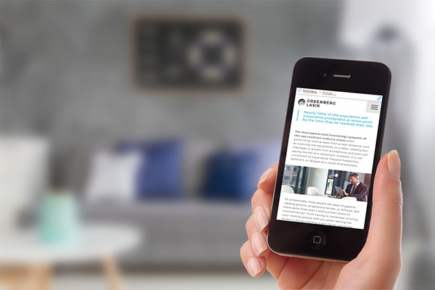 A right hand holding a phone with Greenberg LASIK website on the screen and a living room backdrop with couch and pillows. The phone is in focus while the backdrop is out of focus.